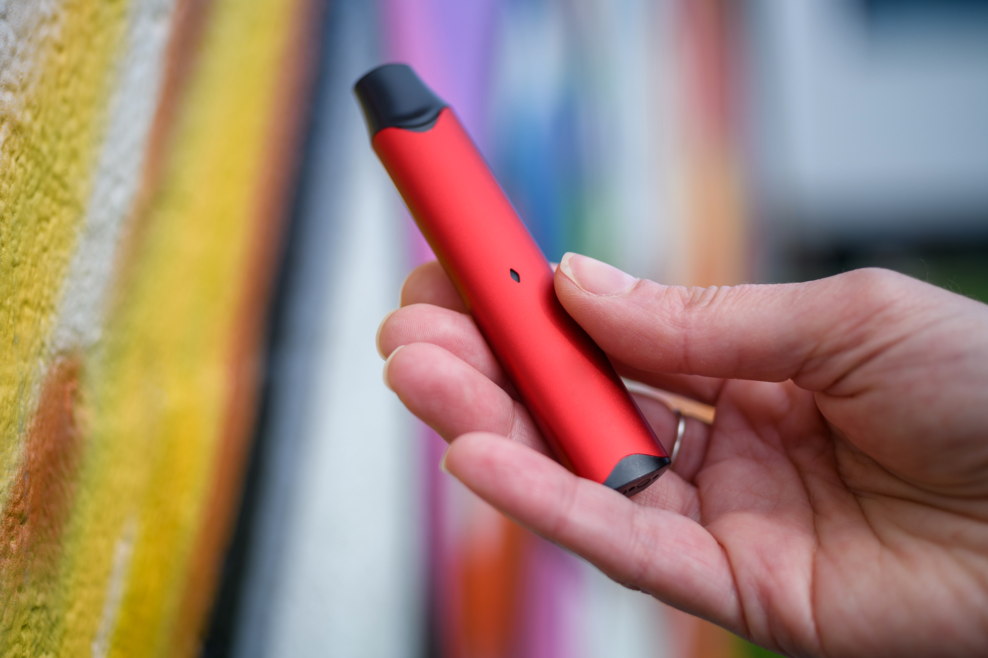 Close-up of person holding red vapor pen next to graffitied wall.